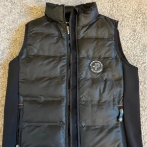 Svart dunväst från Pelle P - Säljer en snygg svart dunväst från Pelle P i storlek 140. Västen har en dragkedja framtill och en stilren design med logga på bröstet. Perfekt för kyliga dagar och passar bra till både vardag och utomhusaktiviteter. Håll dig varm och trendig! 🖤