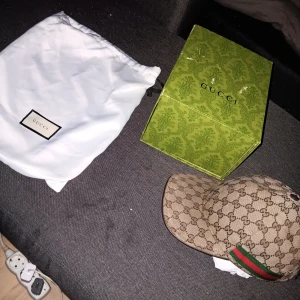 Beige keps från Gucci - Snygg beige keps från Gucci med det klassiska GG-mönstret. Kepsen har en grön och röd randdetalj på sidan. Perfekt för att ge en lyxig touch till din outfit. Box å dustbag fås med 
