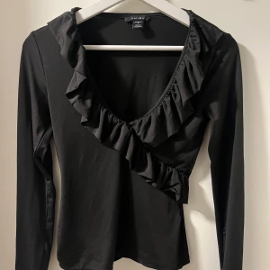 Svart långärmad topp med volang - Snygg svart långärmad topp från Amisu i storlek M. Den har en elegant volangdetalj som går diagonalt över bröstet och en djup v-ringning. Perfekt för både vardag och fest! 🖤