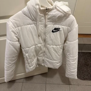 Vit pufferjacka från Nike - Säljer en snygg vit pufferjacka från Nike. Jackan har en dragkedja framtill och en praktisk huva.