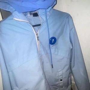 Trapstar Ljusblå vindjacka med huva - Säljer en ljusblå vindjacka med huva och dragkedja framtill. Jackan har en dekorativ knappdetalj på bröstet och justerbara snören vid huvan. Perfekt för blåsiga dagar!