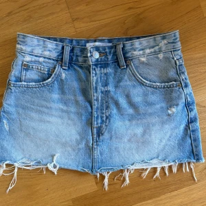 Blå jeanskjol - Säljer en cool blå jeanskjol med slitna detaljer och fransig kant. Perfekt för sommaren och ger en avslappnad look. Kjolen har en knapp och dragkedja framtill samt klassiska fem fickor. Passar perfekt till en t-shirt eller en söt topp!