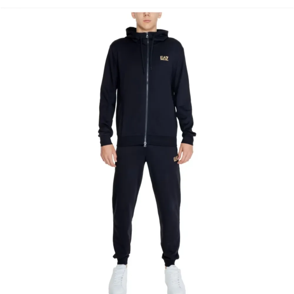 Säljer en stilren svart tracksuit från EA7. Den har en dragkedja framtill och är perfekt för både träning och vardag. Hoodien har långa ärmar och en bekväm passform. EA7-loggan syns diskret på bröstet. Perfekt för dig som vill ha en snygg och funktionell tröja!. Tröjor & Koftor.