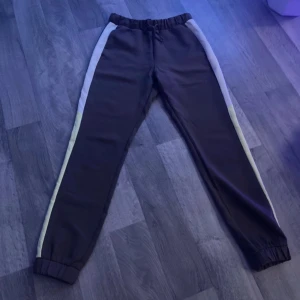 Svarta mjukisbyxor med vita ränder - Säljer ett par svarta mjukisbyxor med vita ränder längs sidorna. De har en elastisk midja med snörning för justerbar passform. Perfekta för en avslappnad dag hemma eller lätt träning. Superbekväma och stilrena!