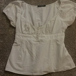 Vit blus från Brandy Melville - Säljer en söt vit blus från Brandy Melville. Den har korta puffärmar och en v-ringad design med knappar framtill. Perfekt för sommardagar eller en avslappnad vårlook. Blusen är i bra skick och passar till både jeans och kjol.