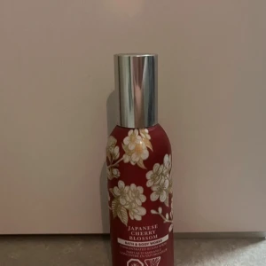 Japanese Cherry Blossom Body Mist - Säljer en elegant rumsspray från Bath & Body Works med doften Japanese Cherry Blossom. Flaskan är röd med vackra vita blommor och har en silverfärgad kork. Perfekt för att fräscha upp sig med en blommig och söt doft.