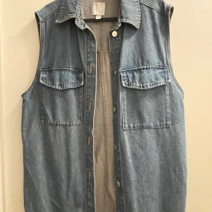 Jeansväst från H&M - Säljer en jeansväst från H&M i storlek S. Västen har en klassisk krage och knappar framtill. Perfekt för lager-på-lager-stil och passar både till vardags och fest. Två bröstfickor med lock ger en extra touch. Skön och avslappnad passform.