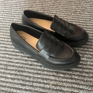 Svarta loafers i skinn - Snygga svarta loafers i skinn, perfekta för både vardag och fest. De har en klassisk design med en bred rem över foten och en robust sula för extra komfort. Skorna är i nyskick och redo att användas direkt. Passar perfekt till både jeans och klänning!