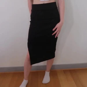 Svart kjol med asymmetrisk slits - Snygg svart kjol med en cool asymmetrisk slits. Perfekt för både vardag och fest! Kjolen är tight och har en stilren design som passar till allt. Ett måste i garderoben för den som gillar att mixa och matcha.