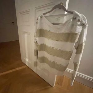 tjocktröja - Säljer en snygg vit och guld randig tröja från Holly & Whyte by Lindex. Den är stickad och har långa ärmar med dekorativa knappar på axlarna.