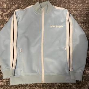 Säljer en snygg ljusblå track jacket från Palm Angels. Jackan har en vit dragkedja och vita ränder längs ärmarna. Den är gjord i 100% polyester och har en normal passform. Perfekt för alla årstider!