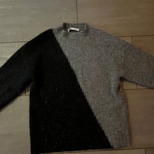 Grå och svart stickad tröja - Säljer en supermysig stickad tröja i grått och svart. Den har en cool diagonal design som verkligen sticker ut. Perfekt för höst och vinter när du vill hålla dig varm och stilren. Tröjan har långa ärmar och en normal passform. Perfekt för både vardag och mysiga hemmakvällar!