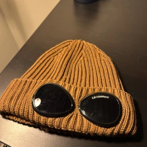 Brun ribbad mössa från C.P. Company - Säljer en snygg brun ribbad mössa från C.P. Company. Den har coola svarta glasögondetaljer framtill som ger en unik look. Perfekt för att hålla sig varm och stilren under höst och vinter. Passar både till vardags och lite mer avslappnade tillfällen.