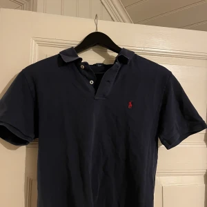 Mörkblå pikétröja från Polo Ralph Lauren - Säljer en klassisk mörkblå pikétröja från Polo Ralph Lauren i 100% bomull. Tröjan har en broderad röd logga på bröstet och korta ärmar. Perfekt för en stilren och avslappnad look. Passar både till vardags och fest! Tröjan är i fint skick och storlek XL i barnstolek, vilket motsvarar ungefär S/M i Herr. Jag är 184 och den har riktigt skön och snygg passform på mig. Fler bilder kan ordnas.