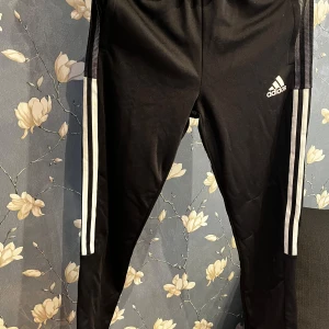 Svarta träningsbyxor från Adidas - Säljer ett par svarta träningsbyxor från Adidas i mycket bra skick. De har de klassiska tre vita ränderna längs sidorna och en elastisk midja för bästa passform. Perfekta för träning eller en avslappnad dag. Adidas-loggan syns tydligt på framsidan. Passar perfekt för både sport och vardag!