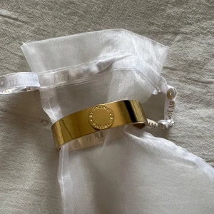 Guldarmband från By Jolima - Snyggt och elegant guldarmband från By Jolima. Armbandet har en stilren design med en rund platta som pryds av märkets logotyp. Perfekt accessoar för både vardag och fest. Kommer i en fin vit organzapåse med band. Nypris 649kr