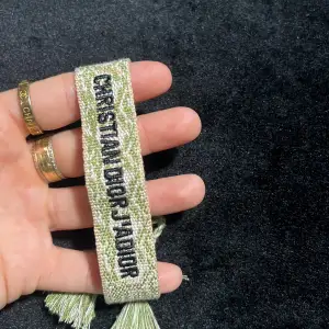 Snyggt grönt armband från Christian Dior med text i svart. Armbandet har en bohemisk stil med fransar i ändarna. Perfekt accessoar för att ge din outfit en lyxig touch. Passar både till vardags och fest! ej ekta