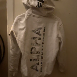 Vit hoodie från Alpha Industries - Säljer en snygg vit hoodie från Alpha Industries. Den har en stor logga på ryggen och en praktisk ficka på ärmen. Perfekt för kyliga dagar och har en skön passform med känguruficka fram. Huvan har dragsko för justering. Passar både till vardags och chill.