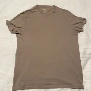 Säljer en enkel och stilren beige t-shirt i bomull. Den har en normal passform och korta ärmar, perfekt för vardagsbruk. T-shirten är i mycket bra skick och passar till alla tillfällen. Skön och lätt att matcha med andra plagg!