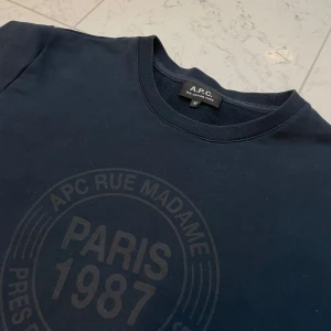 Mörkblå tröja från A.P.C. - Säljer en stilren mörkblå tröja från A.P.C. med tryck 'Paris 1987' på framsidan. Tröjan har en normal passform och är perfekt för både vardag och avslappnade tillfällen. Den är i mycket bra skick och gjord av mjukt bomullsmaterial.