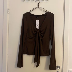 Brun långärmad topp från Gina Tricot - Säljer en snygg brun långärmad topp från Gina Tricot i storlek M. Toppen har en knytning framtill som ger en fin detalj. Den är i nytt skick med prislappen kvar. Perfekt för både vardag och fest! Säljer även en i vit