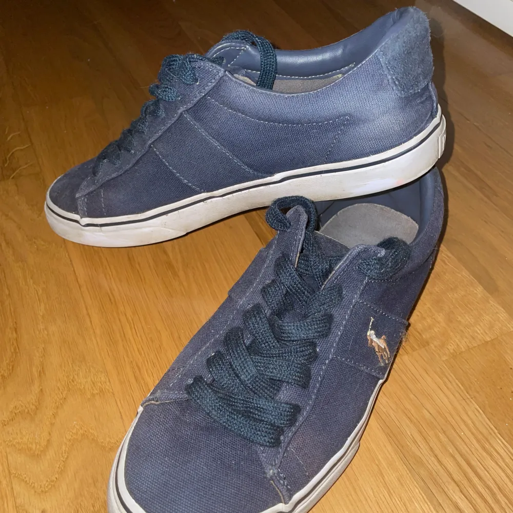 Säljer ett par mörkblå sneakers från Polo i bra skick. Skorna har en klassisk design med snörning och en liten logga på sidan. Perfekta för vardagsbruk och passar till det mesta. Sulan är vit och ger en snygg kontrast till det mörkblå tyget.. Skor.