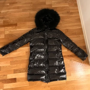 Gloss Jacket - Storlek:  M Glansig jacka med äkta päls. Fodrad och vattenavvisande. Pälsen är avtagbar. Skick: Använd/Ny Material: 100% Polyester Fyllning: 90% duck down  Luva: 100% Raccoon Fur