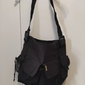 Svart axelväska med fickor - Säljer en praktisk svart axelväska/skötväska med flera fickor och justerbar axelrem. Väskan har en dragkedja upptill och är perfekt för vardagsbruk. Man kan använda som axelväska eller fästa på barnvagn/rullstol. Den har också en nätficka på sidan för extra förvaring. Perfekt för dig som behöver en rymlig och funktionell väska!