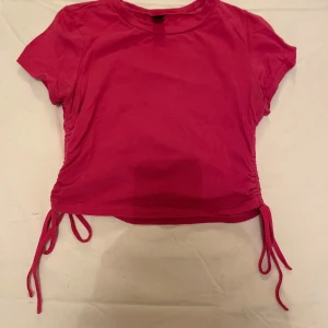 Rosa kortärmad topp från Shein - Säljer en söt rosa topp från Shein i storlek M. Den är kortärmad och har knytband på sidorna för en justerbar passform. Perfekt för sommardagar eller en avslappnad utekväll. Toppen är i bra skick och redo för en ny ägare!