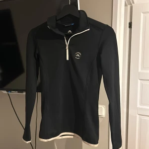 Svart långärmad tröja från J.Lindeberg - Snygg svart långärmad tröja från J.Lindeberg med dragkedja vid halsen. Perfekt för kyliga dagar eller som ett lager under jackan. Den har en stilren design med logotyp på bröstet och ryggen. Passar både till vardags och sportiga aktiviteter.