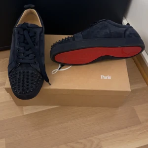 Mörkblåa sneakers med nitar från Christian Louboutin - Säljer ett par svarta sneakers från Christian Louboutin i mocka med coola nitar på tån och sidorna. Skorna har den ikoniska röda sulan och kommer med extra skosnören. Perfekta för att ge din outfit en edgy touch!