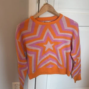 Färgglad stickad tröja med stjärnmönster - Säljer en supercool och färgglad stickad tröja med ett stort stjärnmönster i orange, rosa och lila. Tröjan har en normal passform och långa ärmar, perfekt för att liva upp vilken outfit som helst. Den är i nyskick och redo för nya äventyr!