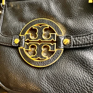 Svart skinnväska med gulddetaljer - Säljer en snygg svart skinnväska från Tory Burch. Väskan har ett stort guldfärgat emblem på framsidan och matchande gulddetaljer på handtagen. Insidan är fodrad med ett ljust tyg med logotypmönster och har en praktisk dragkedja. Perfekt för både vardag och fest!