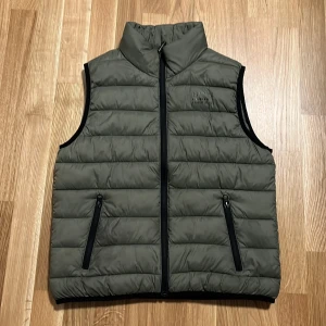 Grön quiltad väst med dragkedja - Säljer en snygg grön quiltad väst med dragkedja framtill. Västen har två praktiska fickor med dragkedjor och en hög krage för extra värme. Perfekt för lager-på-lager under höst och vår!