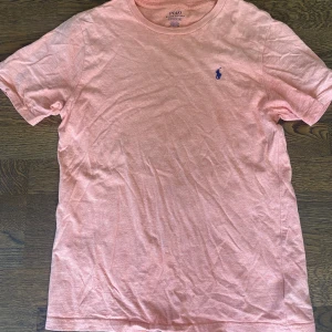 Polo Ralph Lauren T-Shirr - En väldigt fin Polo Ralph Lauren tröja. Den är i fint skick och sparsamt använd i ca 1 månad. Pris 250 kr. Svår att beskriva i färgen men typ korall/rosa.