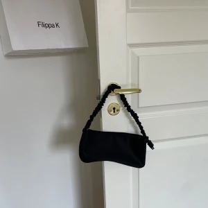 Svart handväska - Säljer en stilren svart handväska. Väskan har en snygg, vriden axelrem och stängs med dragkedja. Perfekt för både vardag och fest!