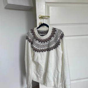 Vit stickad tröja med mönster - Säljer en superfin vit stickad tröja med ett snyggt mönster i brunt och grått runt halsen. Tröjan är perfekt för kyliga dagar och har långa ärmar. Den är i bra skick och redo för en ny ägare!