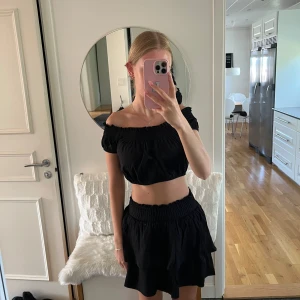 Svart offshoulder topp - Säljer en snygg svart offshoulder topp med matchande kjol som är perfekt för sommaren. Toppen är croppad och plaggen har en elastisk passform som gör de både bekväma och stilrena. Perfekt för en dag på stan eller en sommarkväll med vänner.
