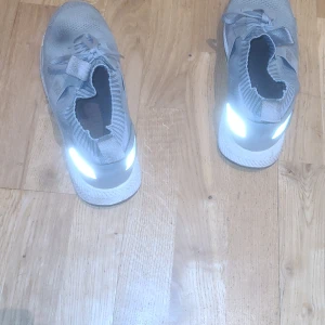 Grå sneakers med stickad design - Säljer ett par grå sneakers med en snygg stickad design. Skorna har snörning och en bekväm passform, perfekt för vardagsbruk. De har en reflekterande detalj på hälen som ger en cool effekt i ljus.