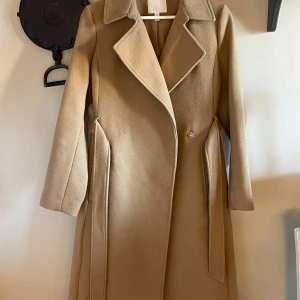Beige kappa från H&M - Säljer en stilren beige kappa från H&M i storlek S. Den har en klassisk design med knappar och ett bälte i midjan för en snygg passform. Perfekt för höst och vår! Materialet är en blandning av polyester och akryl, vilket gör den både varm och hållbar. Ett måste i garderoben för en chic look!