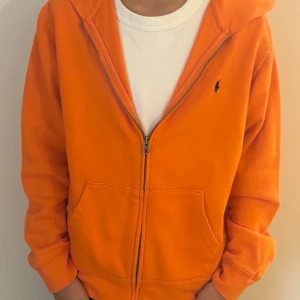 Orange huvtröja från Ralph Lauren - Säljer en snygg orange huvtröja från Ralph Lauren i mycket bra skick. Den har en dragkedja framtill och en klassisk logga på bröstet. Perfekt för höst och vår, med långa ärmar och en skön huva. Passar både till vardags och chillkvällar. Storlek L i barn inte så jätte bra skick ungefär 5/10 skick.