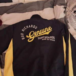 Svart och gul jacka från Bud Richards - Säljer en cool svart och gul jacka från Jack&Jones och har en text med Bud Richards med texten 'Genuine Surfboards Made in Hawaii' på ryggen. Den har en dragkedja fram och nummer 3 på bröstet. Perfekt för en avslappnad stil eller surfvibbar. Jackan är långärmad och i bra skick. Passar perfekt för vår och höst!