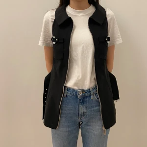 Svart väst från Karl Lagerfeld - Snygg svart väst från Karl Lagerfeld med dragkedja och bälte i midjan. Västen har två fickor med knappar framtill och är perfekt för en stilren look. Passar bra till både vardag och fest.