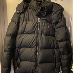 Svart dunjacka från Polo Ralph Lauren - Säljer en svart dunjacka från Polo Ralph Lauren i mycket bra skick. Jackan har en huva och dragkedja framtill, nypris ligger på runt 5500 kronor. Inget kvitto