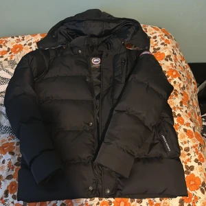 Svart dunjacka från Canada Goose - Säljer en svart dunjacka från Canada Goose i storlek S. Jackan är perfekt för kalla vinterdagar och har en klassisk pufferstil med långärmade ärmar. Den har en dragkedja och knappar framtill samt en broderad logga på ärmen. Supervarm och stilren!