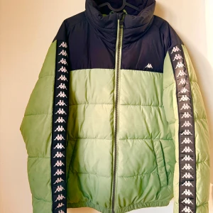 Grön och svart pufferjacka från Kappa - Säljer en snygg grön och svart pufferjacka från Kappa. Jackan har en dragkedja framtill och ikoniska Kappa-loggor längs ärmarna. Perfekt för kyliga dagar med sin quiltade design som håller värmen. Den är i mycket bra skick och passar perfekt för höst och vinter.