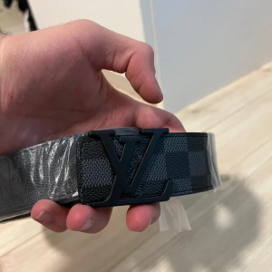 Rutig bälte med svart spänne - Säljer ett snyggt bälte med ett klassiskt rutigt mönster i svart och grått. Bältet har ett stilrent svart spänne och är i nytt skick, fortfarande inplastat. Perfekt för att ge en touch av stil till vilken outfit som helst!