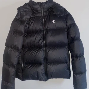 Svart dunjacka från Calvin Klein - Säljer en snygg svart dunjacka från Calvin Klein. Den är perfekt för kalla dagar med sin pufferstil och har en dragkedja framtill. Jackan har långa ärmar och en liten CK-logga på bröstet. Den är i mycket bra skick och passar perfekt för både höst och vinter. (Hör av er vid fler bilder)💞