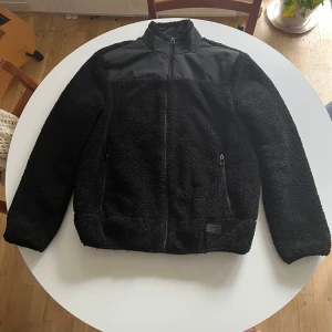 Svart fleecejacka från Sams Black - Mysig svart fleecejacka från Sams Black i storlek M. Perfekt för höst och vinter med dragkedja och hög krage. Jackan har en cool kontrastdetalj i slätare material över axlarna och bröstet. Den är långärmad och har praktiska fickor med dragkedja. Superbekväm och stilren för alla tillfällen!
