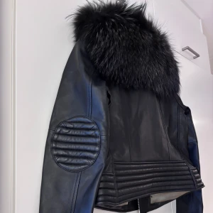 Svart skinnjacka med pälsdetalj  - Säljer en snygg svart skinnjacka från Custommade by Numbers med en fluffig pälsdetalj på kragen. Jackan har en cool bikerstil med dragkedjor och quiltade detaljer på ärmarna och nederkanten. Perfekt för höst och vinter, och ger en edgy look till vilken outfit som helst. Passar bra till både vardag och fest!